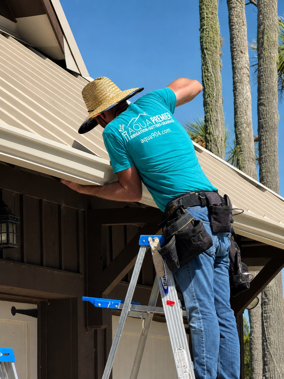 Aqua Premier technician in a branded shirt installing a seamless aluminum gutter from a ladder in Ponte Vedra Beach, Florida